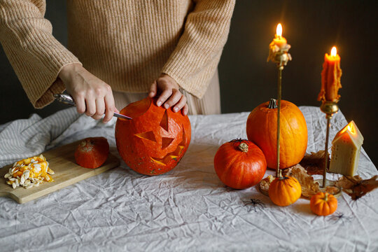 Happy Halloween. Woman carving halloween pumpkin on table with scary decorations and burning candles on dark moody background. Space for text. Making jack o lantern