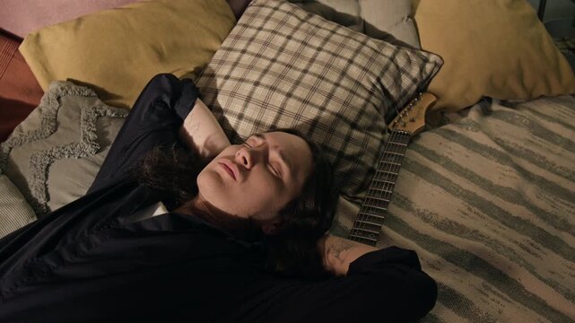 Tired young man lying on bed with eyes closed next to electric guitar, resting. Footage for mental health campaigns, creative burnout stories, or music lifestyle advertisements