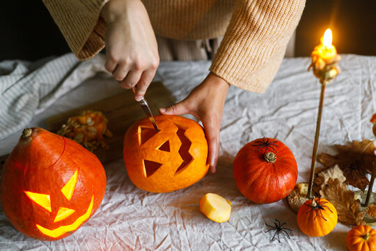 Happy Halloween. Woman carving halloween pumpkin on table with scary decorations and burning candles on dark moody background. Space for text. Making jack o lantern