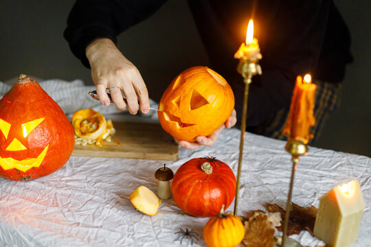 Carving halloween pumpkin. Hands holding pumpkin and knife on table with burning candles and spiders on moody background. Making jack o lantern, halloween preparation and decorations