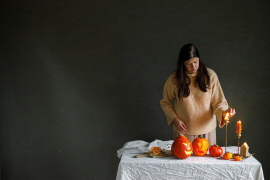 Making jack o lantern. Woman hands lighing up candles and carving pumpkins on table with spiders on moody background. Halloween preparation and decorations