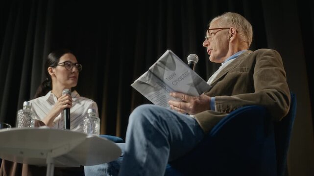 Senior Caucasian man holding book and speaking into microphone during presentation while adult woman listens. Footage is ideal for promoting literary events or educational seminars, low angle