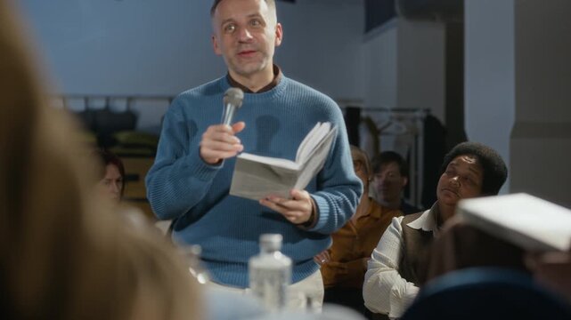 Middle-aged Caucasian man raising hand, standing up, and speaking into microphone while holding book at presentation. Clip is perfect for educational events or community workshop promotions