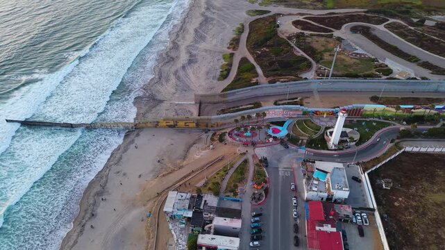 A striking high-angle drone shot framing the international border wall, the historic lighthouse, and the coastal park of Playas de Tijuana.