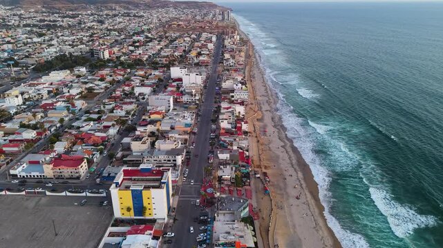 An elevated aerial drone perspective showcasing the steel international border wall as it snakes across the steep, undulating terrain of the coastal hills near the Pacific shoreline.