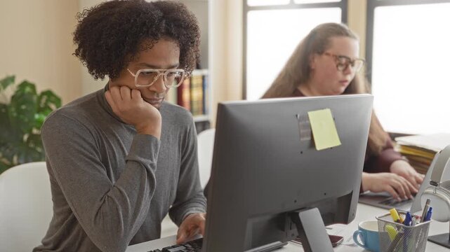 Man at computer yawning with hand covering mouth, seated at desk with monitor and sticky note while coworker types at laptop in a building office with bookshelf and plant; fatigue.