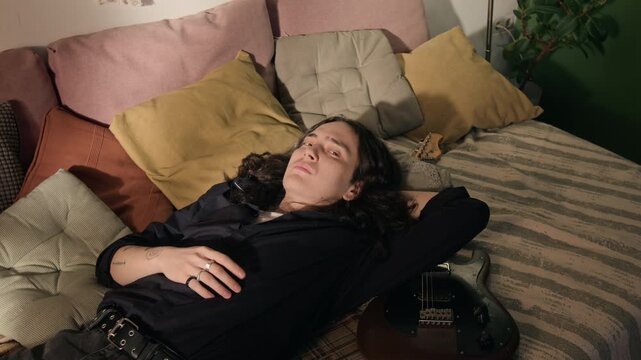 Young man lying on bed next to electric guitar, opening eyes and looking at camera. Footage for mental health awareness, creative block, or youth lifestyle campaigns