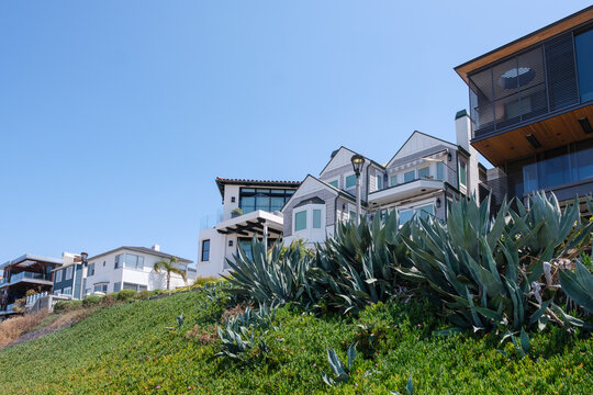 Luxury beachfront houses and modern residential architecture along The Strand, framed by green plants and agaves under a blue sky in Manhattan Beach, Los Angeles, California.