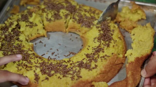 Close-up shot of a person using a metal spoon to slice and serve a golden ring-shaped cake covered in chocolate sprinkles on a metal tray.