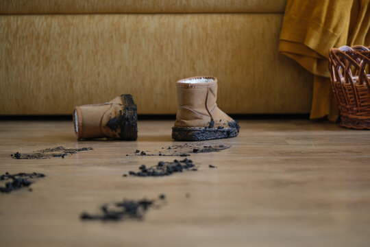 A pair of muddy children's winter boots left on a wooden floor after outdoor play, with scattered clumps of dirt and mud creating a messy trail indoors. Home care and cleaning routine.