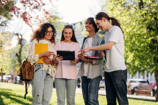 Group of four young women collaborating outdoors in a park, holding notebooks and a tablet, surrounded by greenery and trees on a sunny day