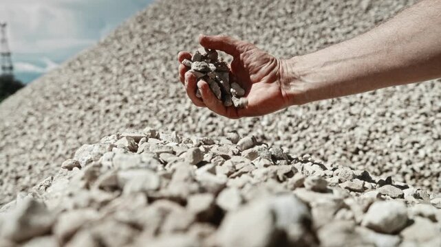Worker hand releasing crushed stone over gravel mound during quarry quality inspection in outdoor industrial setting with rough texture, dust, daylight, and material sampling