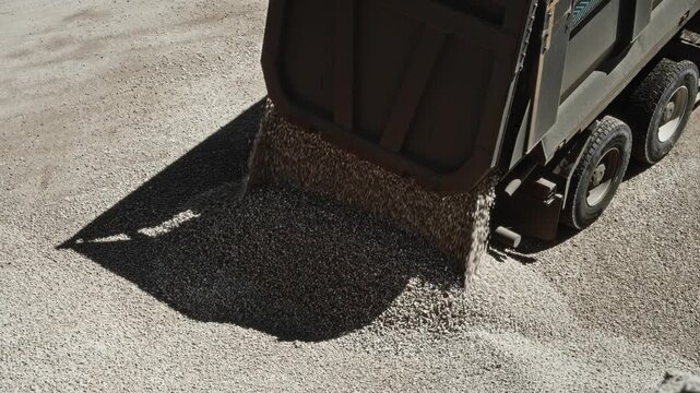 Dump truck unloading crushed stone onto compacted gravel at construction site, rear bed tilted and tailgate open as material falls into growing pile in bright sunlight