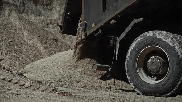 Rear dump truck unloading crushed stone onto large gravel pile at outdoor aggregate yard, low angle view showing tilted bed, open tailgate, heavy load discharge and dust