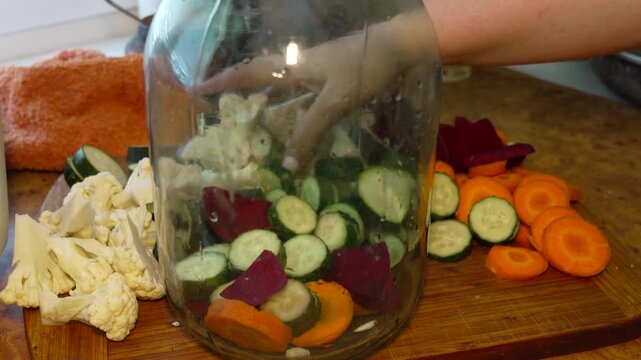 Vegetables, canning, glass, woman adds fresh cauliflower florets into large glass jar for pickling process with organic cucumber beet and carrot slices