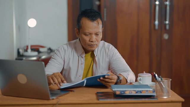 Focus and relaxed of Asian man in casual shirt reading a notebook in home office desk. Learning, analyzing, literacy and education concept.