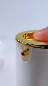 Front close up of a finger pressing and pumping the cap of a beauty product bottle. Gold and white elegant packaging. Skincare or cosmetic product being dispensed for daily use.