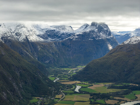 Aerial view of the Troll Wall with snow-capped peaks, lush green valley, winding river, and agricultural fields in Rauma Municipality, More og Romsdal, Norway.