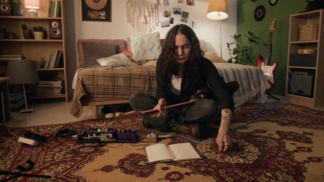 Young man sitting on floor, playing electric guitar and writing song lyrics in notebook. Footage for music education, creative hobby, or artistic lifestyle ads