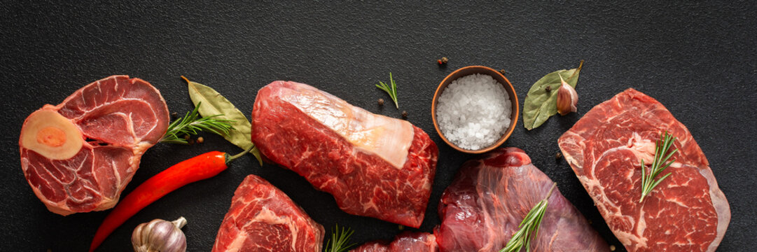 Various cuts of beef steak are shown with herbs, spices, and oil on a dark surface. Perfect for those looking to prepare or grill different beef dishes at home, banner