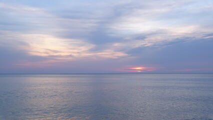 Fototapeta premium Serene pastel sky with soft clouds over a calm ocean at dawn. A gentle and dreamy dawn scenery showing pink and purple sky colors with a tiny glimpse of the sun rising over a peaceful, flat sea.