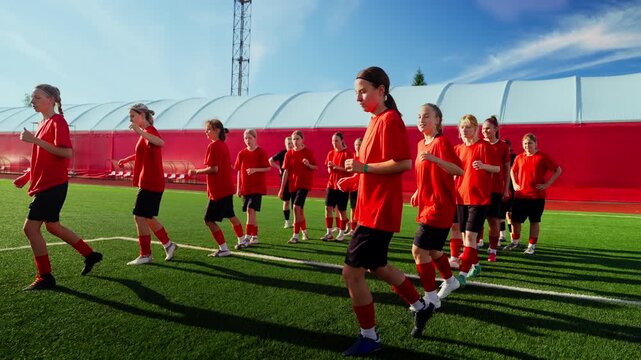 Youth womens soccer team warming up together on green artificial turf field during outdoor training session under clear blue sky with coordinated group drills