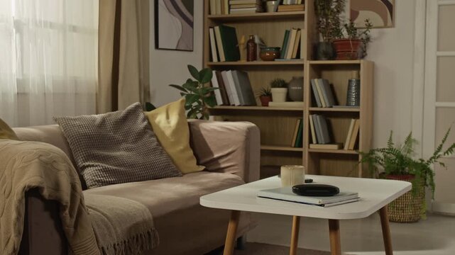No people shot of empty modern living room featuring beige sofa with pillows, coffee table, and wooden bookshelf. For real estate, interior design, or home decor advertising