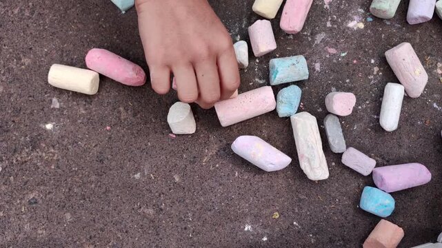 Child reaching for colorful sidewalk chalk scattered on pavement, outdoor creativity, playful learning, summer childhood, artistic activity.