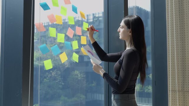 Creative professional businesswoman writing ideas strategy marketing and tasks on colorful sticky notes on glass wall in modern office