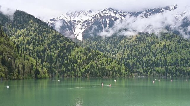 The emerald green waters of Lake Ritsa with small boats, framed by dense wooded hillsides and majestic snowy mountains, Abkhazia
