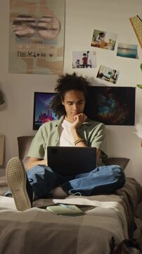 Vertical full video of Black male teenager sitting on bed, using laptop for studying and thinking. Footage is perfect for marketing online education, e-learning, and student lifestyle