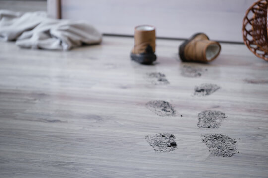 Footprints lead across a wooden floor next to old boots and a rolled blanket on the gray floor. The scene shows a relaxed indoor space with personal items scattered around.