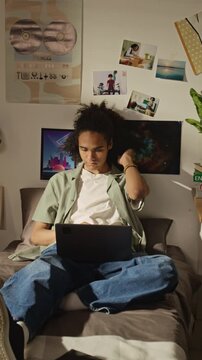 Vertical full shot of Black teenager sitting on bed, typing on laptop and thinking while studying in bedroom. Footage represents online education, remote learning, and student lifestyle concepts