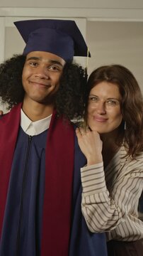 Vertical slowmo portrait of African American male teenager in graduation gown smiling with Caucasian middle aged mother. For university enrollment campaigns and family financial planning ads