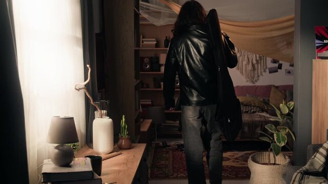 Young musician entering room, putting down guitar case, taking off leather jacket, and collapsing onto bed to rest. Perfect for lifestyle commercials or mental health campaigns