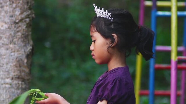 Little girl in tiara holding praying mantis outdoors, curious child exploring nature, candid summer moment in park with colorful playground background