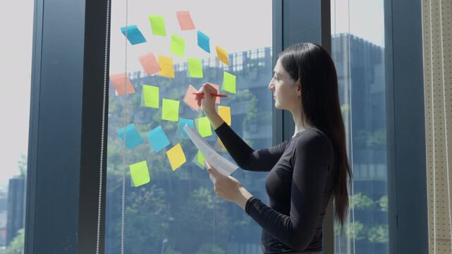 Creative professional businesswoman writing ideas strategy marketing and tasks on colorful sticky notes on glass wall in modern office