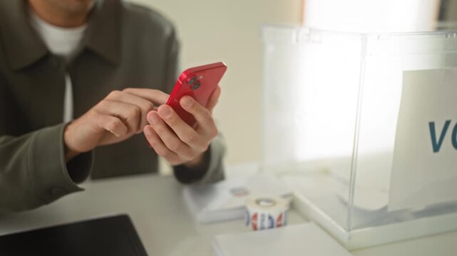 Man taps red smartphone with index finger at polling table in building near ballot box and us flag; civic duty determined.