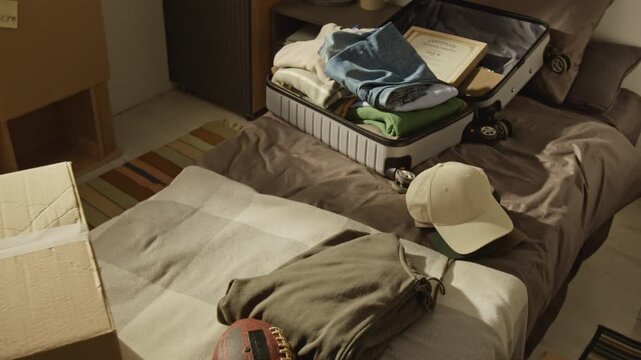No people shot of open suitcase lying on bed filled with folded clothes and graduation certificate next to cardboard moving boxes. Footage is ideal for ads about college relocation or student housing