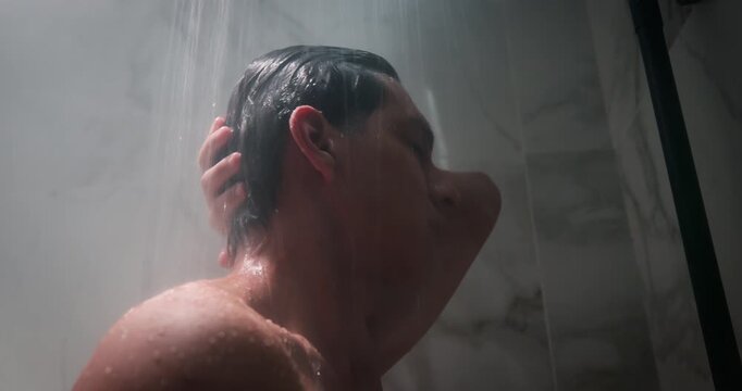 Man standing under shower in bathroom. Relaxed guy enjoying hot water stream. Daily morning evening hygiene routine, skincare self care concept. Wellness, health. Person use shampoo, gel, conditioner.