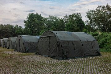 Olive military tents line a green field under a cloudy sky, an army camp during a field exercise. © Victor