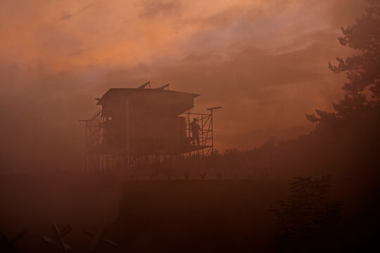 A watchtower silhouette stands in orange smoky haze at dusk, a moody military exercise scene.