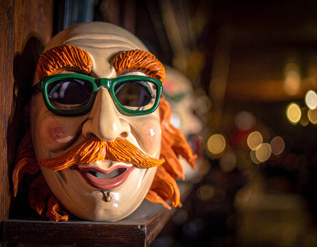 A traditional wax mask with green glasses and orange whiskers used in the Carnival of Binche sits on a shelf.
