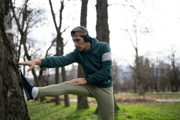 Obraz premium A young man stretches his leg against a tree while wearing headphones and a hoodie. He prepares for an outdoor workout in a quiet park setting.