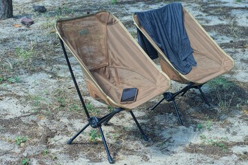 Two brown folding tourist chairs are installed on a sandy glade in a pine forest. There is a smartphone on one chair, and a T-shirt on the other, creating an atmosphere of relaxation during camping  © FEDOR