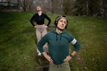 Naklejka premium Man wearing headphones stands with hands on hips and eyes closed in a grassy park. A woman in the background stretches with hands on hips on a step pathway.