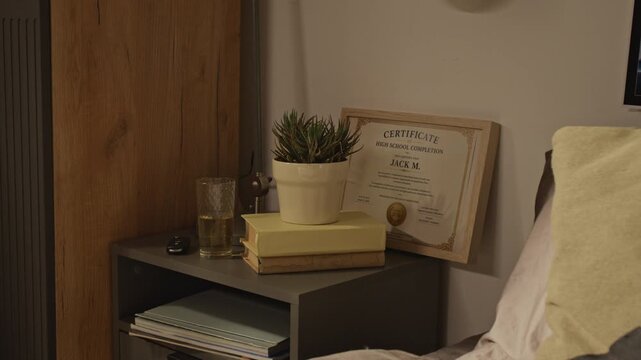 No people close up shot of high school graduation certificate in frame standing on bedside table next to books and plant. For educational marketing or student achievement campaigns