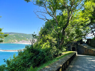 Fototapeta premium Tree shaded stone fort path above turquoise San Sebastian bay in late spring sunlight, Spain