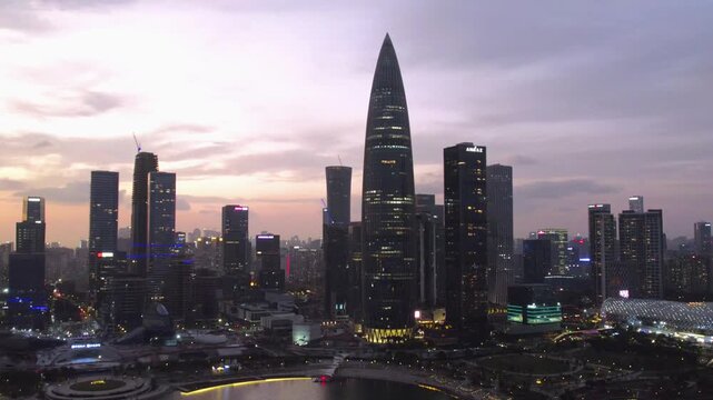 A modern urban skyline with distinctive skyscrapers, their lights on at dusk, reflecting on adjacent water. 