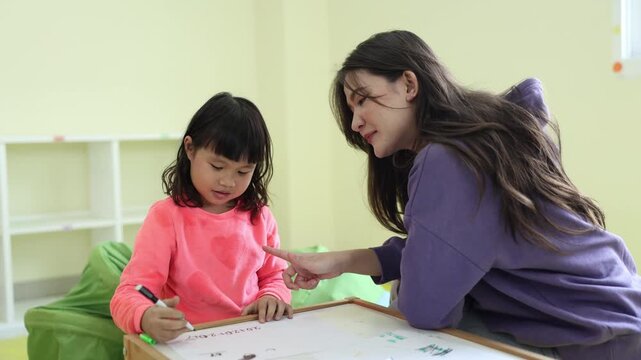 A friendly, smiling educator lovingly helps a young student understand a lesson, guiding them as they write on a whiteboard together in a bright elementary school classroom.